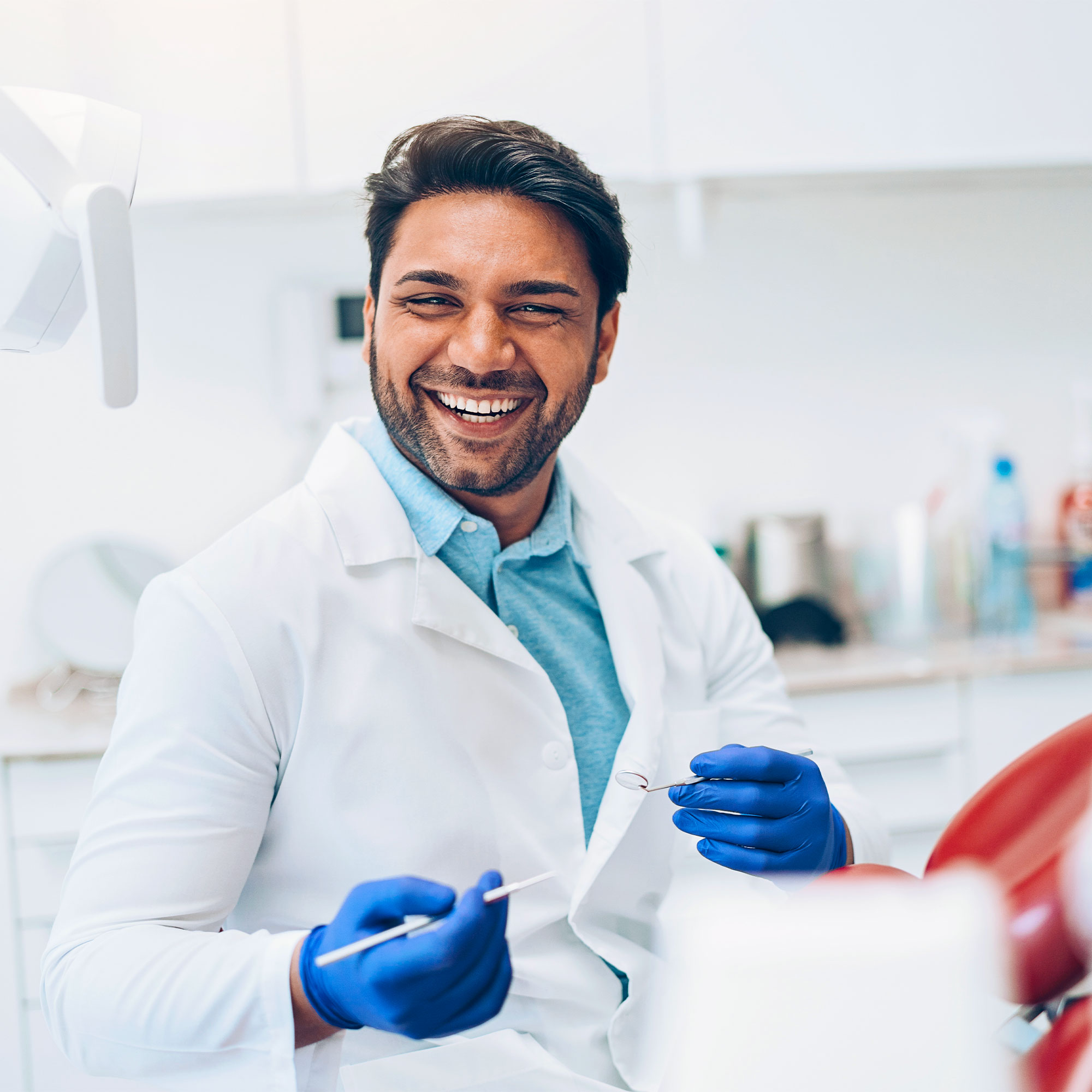 A man in a white lab coat and blue gloves smiles as he sits in his office.